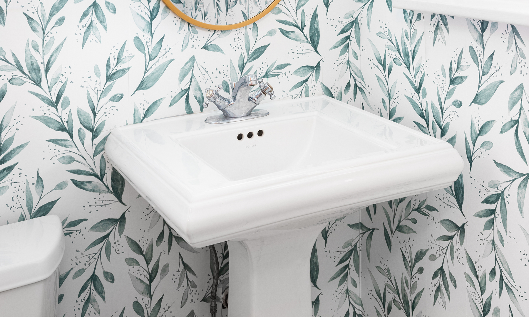 White pedestal sink with chrome faucet in a bathroom with white wallpaper with green leaves on it