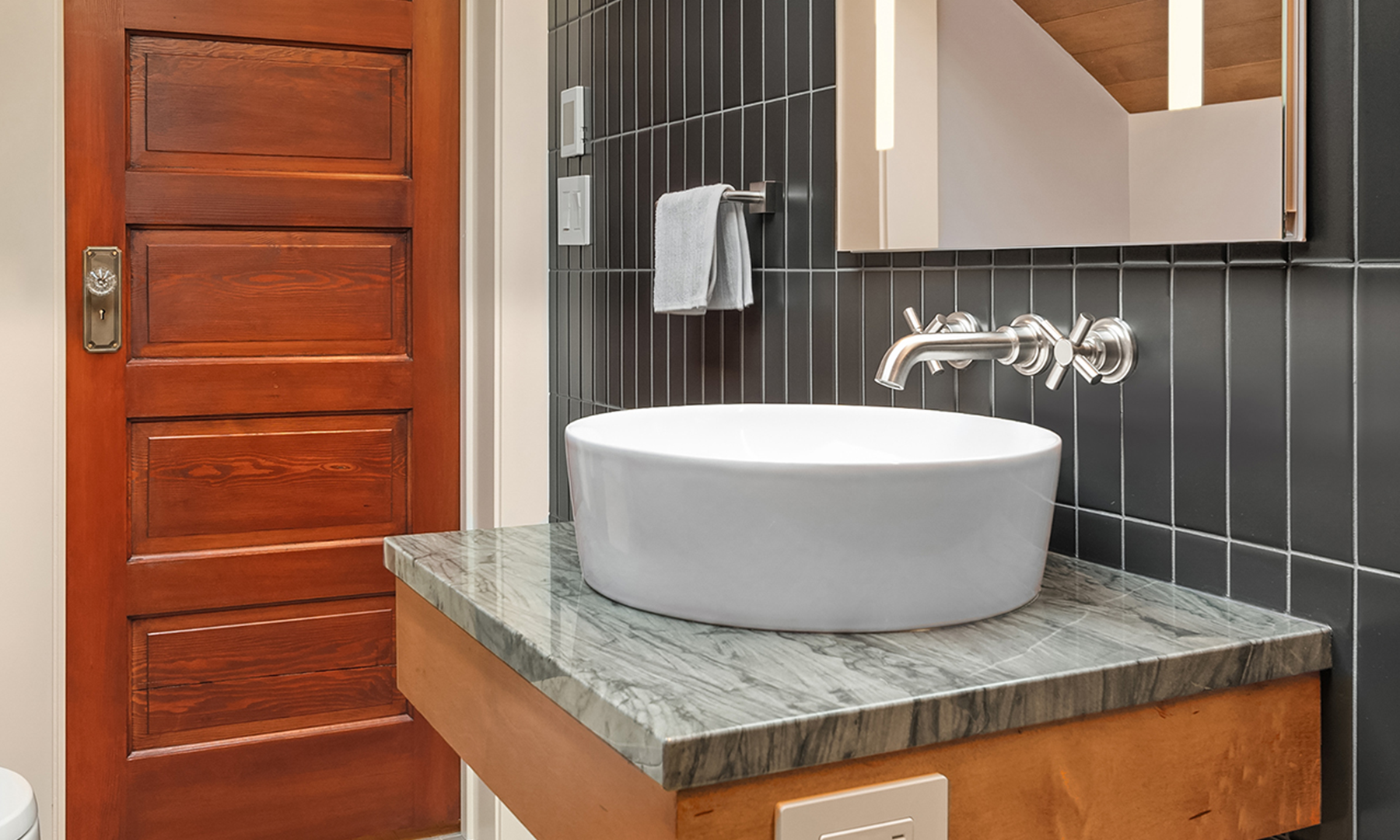 White vessel sink with a chrome wall mounted faucet on a gray granite countertop vanity and black tile wall